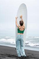 Rear view of woman surfer arms raised holding surfboard vertically, standing on sandy beach, posing photo