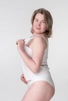 Serious woman in white underwear puts hand on stomach while holding strap of camisole photo