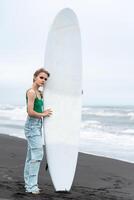 Woman surfer standing on beach, holding surfboard vertically on background of breaking ocean waves photo