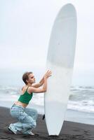 Woman surfer squats and holding surf board vertically on beach against background of ocean waves photo