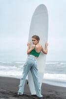 Rear view woman surfer standing on beach, holding surfboard vertically and looking over shoulder photo