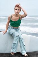 Woman wearing top and blue jeans sitting on surf board lying on sand, on background of sea waves photo