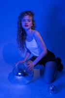 Woman in music style with disco ball partying in nightclub. Woman kneeling in neon colored lights photo