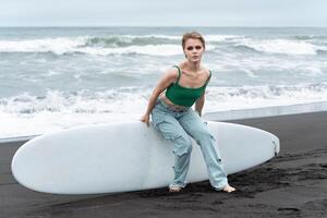 Sensual blonde woman sitting on surf board lying on sand, against backdrop of waves of Pacific Ocean photo