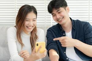 Asian couple smiling and happily use smartphone to online shopping. Husband ready to pay birthday present for beautiful wife. Find accommodation online for honeymoon, App online, book flight tickets. photo