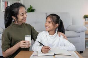 una niña asiática aprende en casa. hacer la tarea con la amable ayuda de la madre, animar para el examen. mamá le pasa un vaso de leche a su hija. niña feliz educación en el hogar. mamá enseña y aconseja educación juntos. foto