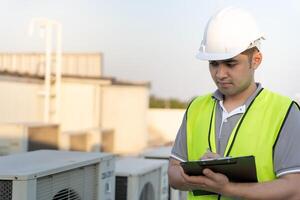 asiático mantenimiento ingeniero trabajos en el techo de fábrica. contratista inspeccionar compresor sistema y planes instalación de aire condición sistemas en construcción. Lista de Verificación, inspector, controlar foto