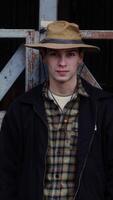 portrait de une Jeune agriculteur avec une sur de soi regard, portant une patiné chapeau et une classique plaid chemise à une rural Grange video