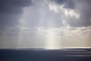 Overcast grey sky with low textured clouds. Sunlight peeks through the clouds and falls on the sea. Background. Horizontal photo