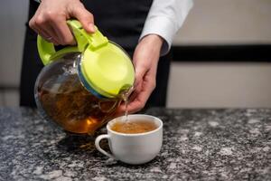 Close-up female hands pour tea from a glass teapot with a green lid into a white mug on marble table photo