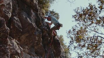 Masculin grimpeur dans action sur une ensoleillé falaise, précisément navigation le rocheux terrain avec une dynamique toile de fond de feuillu des arbres en dessous de une clair bleu ciel video