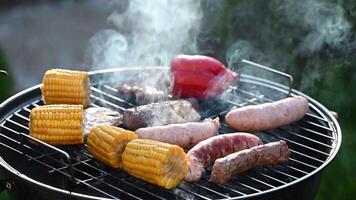 Process of frying and turning fried sausages with corn and red pepper on a round black grill with steam with a landscape in the country video