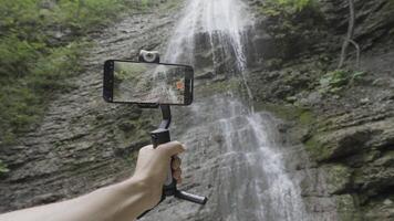 parte inferiore Visualizza di montagne e cascata attraverso il smartphone telecamera. azione. vicino su di mano Tenere un' tripode con fisso telecamera e tiro selvaggio naturale paesaggio. video