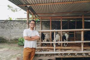 Excited young Asian man standing in front of traditional cage made from wood and bamboo in Indonesia rural area with goat inside photo