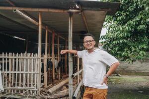 Excited young Asian man standing in front of traditional cage made from wood and bamboo in Indonesia rural area with goat inside photo