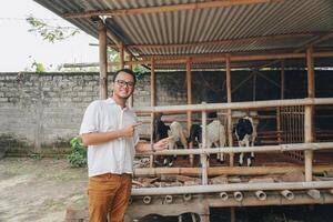 Excited young Asian man standing in front of traditional cage made from wood and bamboo in Indonesia rural area with goat inside photo