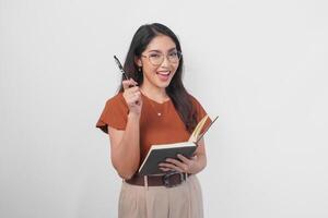 Happy young Asian woman wearing brown shirt and eyeglasses with finding idea gesture while holding book isolated by white background. photo