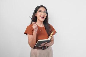 contento joven asiático mujer vistiendo marrón camisa y los anteojos con hallazgo idea gesto mientras participación libro aislado por blanco antecedentes. foto