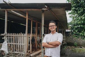 Excited young Asian man standing in front of traditional cage made from wood and bamboo in Indonesia rural area with goat inside photo