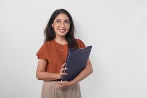 Young Asian woman wearing brown shirt holding a document book report while looking upwards, isolated by white background. photo