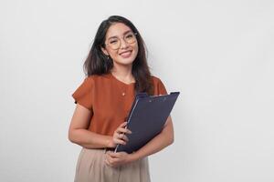 Attractive smiling Asian woman wearing brown shirt and eyeglasses holding document book isolated over white background. photo