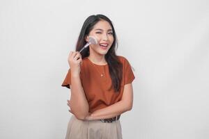 atractivo joven asiático mujer participación maquillaje cepillo a aplicar sonrojo mientras consiguiendo Listo con contento expresión aislado blanco antecedentes. foto