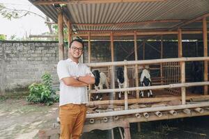 Excited young Asian man standing in front of traditional cage made from wood and bamboo in Indonesia rural area with goat inside photo