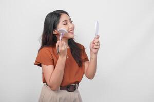 Beautiful young female holding mirror and makeup brush to apply blush using new cosmetics while getting ready isolated white background. photo