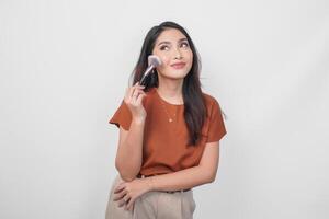 sonriente joven asiático mujer participación maquillaje cepillo a aplicar sonrojo mientras consiguiendo Listo aislado blanco antecedentes. foto