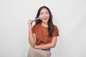 Attractive young Asian woman holding makeup brush to apply blush while getting ready with happy expression isolated white background. photo