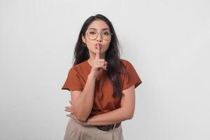 Young Asian woman wearing brown shirt and eyeglass looking to camera while putting a finger in front of lips gesturing stay silent or stay quiet, isolated by white background. photo