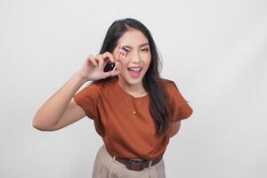 A happy young Asian woman is smiling and holding eyelash curler while wearing brown shirt over white isolated background. photo