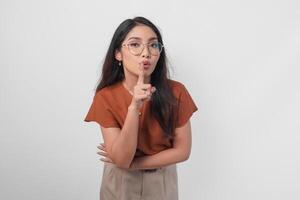 Young Asian woman wearing brown shirt and eyeglass looking to camera while putting a finger in front of lips gesturing stay silent or stay quiet, isolated by white background. photo