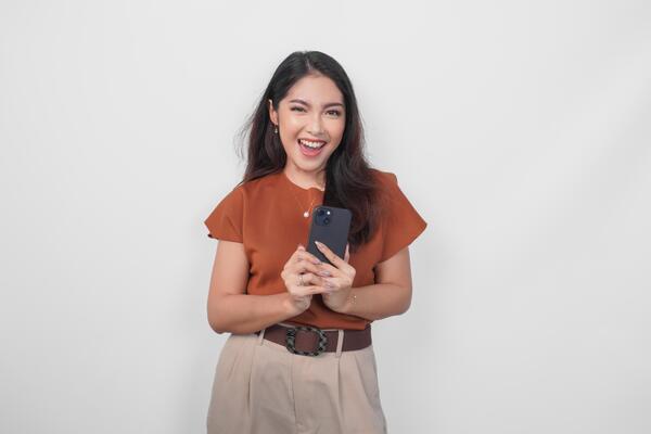 Excited young Asian woman is smiling to the camera and holding her smartphone wearing brown shirt standing over isolated white background.