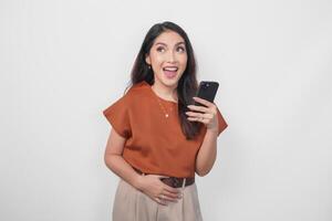 Smiling young woman wearing brown shirt feeling hungry and looking aside while holding smartphone isolated over white background. photo