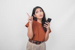 hermosa asiático mujer vistiendo marrón camisa utilizando teléfono inteligente mientras señalando a el Copiar espacio al revés su, aislado por blanco antecedentes. foto