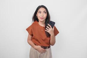 Hungry young woman wearing brown shirt looking aside while holding smartphone isolated over white background. photo