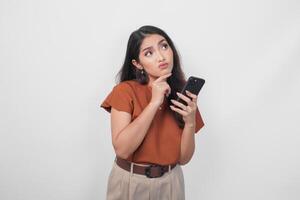 pensativo joven mujer vistiendo marrón camisa mirando aparte mientras participación teléfono inteligente aislado terminado blanco antecedentes. foto