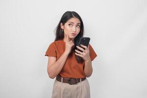 Thoughtful young woman wearing brown shirt looking aside while holding smartphone isolated over white background. photo