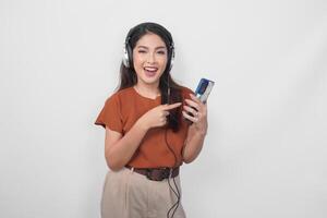 Attractive young Asian woman in brown shirt pointing to the smartphone and listening to music using wireless headphone. photo