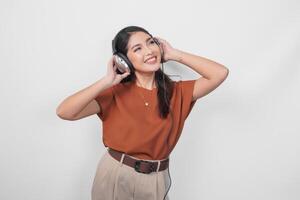 alegre joven asiático mujer en marrón camisa disfrutando y escuchando a música utilizando inalámbrico auricular. foto