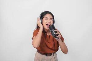 lleno de alegría joven mujer vistiendo marrón camisa y auricular a escucha música y canto a lo largo aislado terminado blanco antecedentes. foto