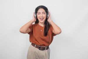 Portrait of a young Asian woman in brown shirt enjoying and listening to music using wireless headphone over isolated white background. photo