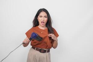 Attractive Asian woman in brown shirt feeling upset while using hairdryer trying to make hairstyle, isolated over white background. photo