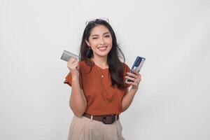 Beautiful young Asian woman wearing brown shirt is smiling while holding her smartphone and credit card over isolated white background. photo