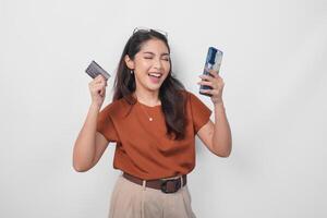 Happy successful Asian woman wearing brown shirt is holding her smartphone and credit card over isolated white background. photo