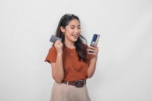 Beautiful young Asian woman wearing brown shirt is smiling while holding her smartphone and credit card over isolated white background. photo