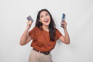 Happy successful Asian woman wearing brown shirt is holding her smartphone and credit card over isolated white background. photo