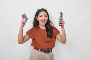 Happy successful Asian woman wearing brown shirt is holding her smartphone and credit card over isolated white background. photo