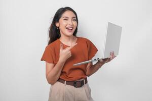 Joyful woman wearing a brown shirt pointing to the laptop she holds, isolated by white background. photo